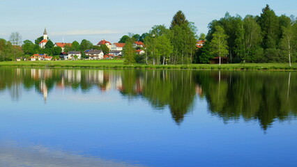Fototapeta na wymiar Blick über stillen Bad Soier See auf Bayersoien mit Spiegelung von Dorf und Wald unter blauem Himmel