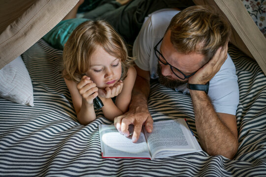 Portrait of a 6 year old boy and his father reading a book in teepee tent. Father and son with flashlight reading book under blanket at home. 