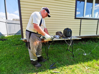 An elderly man cuts metal pipes with a disc grinder. The master works as a grinder in the yard of a country house on a sunny summer day.