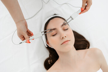 A young girl with a white bandage on her head lies in the cosmetologist's office, view from above. the hands of a female cosmetologist perform a microcurrent facial treatment with the machine. spa