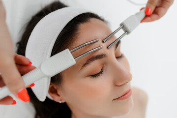 A young girl with a white bandage on her head lies in the cosmetologist's office, view from above. the hands of a female cosmetologist perform a microcurrent facial treatment with the machine. spa