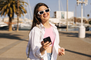Happy young caucasian woman is using modern smart phone spending time outdoors. Brunette wears sunglasses, top and shirt. City life concept