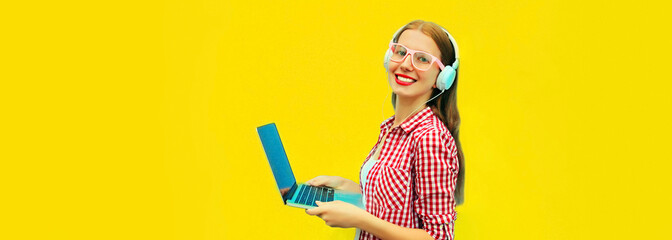Portrait of modern young woman working with laptop listening to music in headphones on yellow background