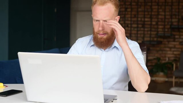 Upset redhead businessman sitting at the desk in front of PC, disappointed and worried male freelancer holding head with hands, has trouble and difficulties, computer error, made a mistake in work. 4k