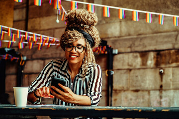 Portrait of young adult black woman with afro hairstyle and eyeglasses texting on smartphone in outdoors coffeehouse shot