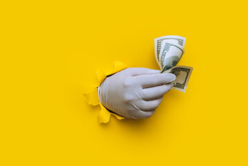 A right doctor holds money (dollars) in white medical glove. Bright yellow torn hole paper background. The concept of treatment fees, bribes, illegal surgery. Copy space.