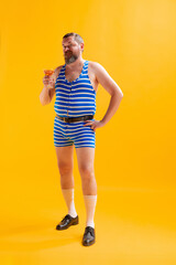 Fototapeta na wymiar Funny seaman, fat cheerful man wearing retro striped swimsuit posing isolated on bright yellow background. Vacation, summer, humor