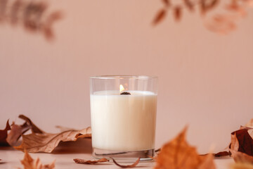 Burning vanilla candle on beige background. Warm aesthetic autumn composition with and dry leaves...
