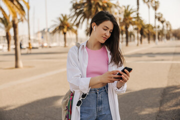 Calm young caucasian woman is rewritten in social networks through smartphone walking around European city on vacation. Brunette wears top, shirt, jeans and bag. Lifestyle concept