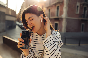 Fototapeta premium Pretty young caucasian woman sings energetically using smartphone as microphone in street. Brunette with closed eyes wears sweatshirt on walk. Summer playful mood concept