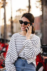 Fototapeta premium Positive young caucasian brunette woman in sunglasses talking on phone walking outdoors. Girl uses wireless device. Happy weekend concept.