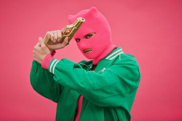 Portrait of bandit woman in pink balaclava crossed her arms with gun in front of her face and...