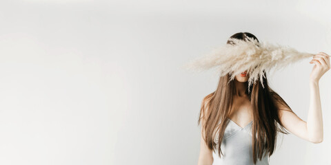 An attractive brunette hides her face behind a fluffy dry branch of a plant on a light gray...