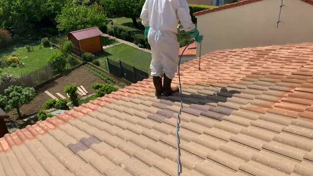 Worker In Protective Suit Spray Painting Roof Tiles Of A House With Waterproofing Paint. high angle