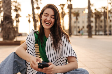 Funny young caucasian girl closing her eyes laughing spending time outdoors with phone. Brunette wears casual clothes. Relaxation concept, technology