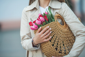 Spring portrait of a young beautiful happy woman 28 years old with long well-groomed hair holds a wicker bag in her hands with a bouquet of tulips on a city street. Stylish model in a trench coat.