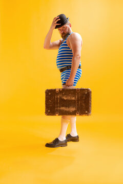 One Funny Beachgoer, Fat Excited Man Wearing Retro Striped Swimsuit And Vintage Bowler Hat Holding Old Suitcase Isolated On Bright Yellow Background. Vacation, Summer, Humor