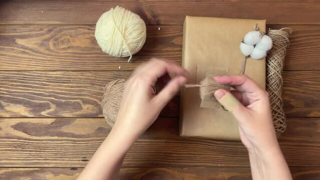 Girl's hands wind brown yarn on piece of cardboard. Handmade crafting decorations of gift for Christmas or Birthday. Box in craft wrapper, twine, cotton and scissors on wooden table. Festive mood.