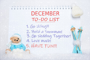 December to-do list. Notepad with to-do list: skiing, making a snowman, sledding, loving, having fun and a toy Teddy bear in blue clothes, blue skis, a white hat on white snow and snowflakes
