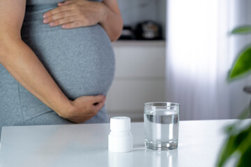 Medicine jar with pills and glass of water on table and pregnant woman with big belly at home interior on background. Health care and prenatal treatment. Vitamins, pills and tablets on pregnancy time. - obrazy, fototapety, plakaty