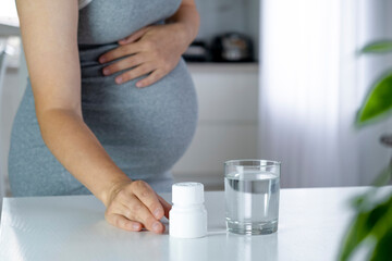 Fototapeta na wymiar Pregnant woman take jar with pills and glass of water at home. Health care and prenatal therapy. Multivitamins, folic acid, Omega-3, anemia. Vitamins, pills and tablets on pregnancy time.