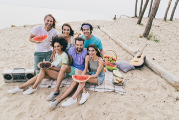 Photo of positive sweet young people wear casual clothes sitting beach eating berries outside countryside