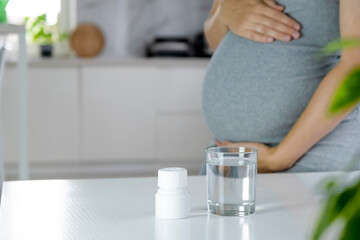 Obraz na płótnie Canvas Medicine jar with pills and glass of water on table and pregnant woman with big belly at home interior on background. Health care and prenatal therapy. Multivitamins, folic acid, Omega-3, anemia.
