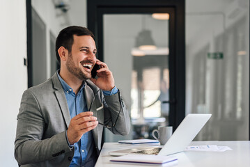 Cheerful entrepreneur talking on cellphone while working on laptop at office