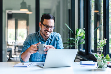 Happy male professional having coffee while working on laptop at home or office - Powered by Adobe