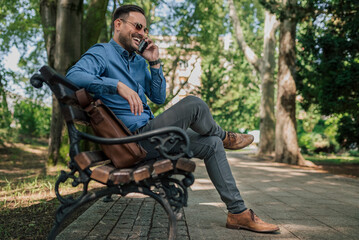 Smiling handsome entrepreneur talking on smart phone while sitting on park bench