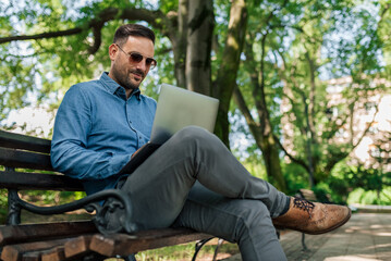 Obraz premium Confident young businessman using laptop while sitting on bench at park