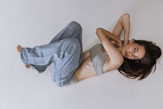 Portrait of young beautiful girl lying on floor, posing in jeans and cotton underwear top isolated over grey studio background. Wellness