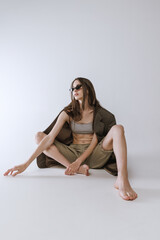 Portrait of beautiful stylish young girl sitting on floor, posing isolated over grey studio background. Trendy outit.