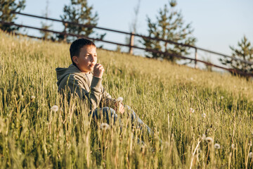 Portrait of happy boy sitting on top of hill in grass field and enjoying beautiful landscape at sunset. Teenager hiker resting in nature. Active lifestyle. Concept of local travel