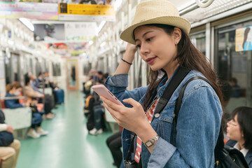 pretty Chinese girl wearing hat using cellphone while riding in a train taking Osaka metro in japan. she stands in the car with other passengers sitting in seats at background