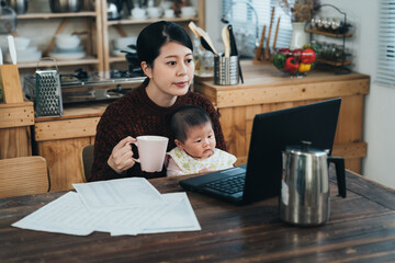 asian career mother working from home is drinking coffee while looking at the laptop monitor with her baby girl sitting in arms at table in the dining room.