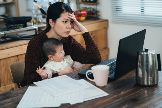 Asian Mom Sitting Holding Her Baby At Desk Is Staring At Computer With A Serious Expression While Working From Home On A Difficult Project In The Dining Room.