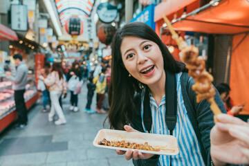 selective focus of happy asian woman giving bbq grilled chicken skewer to camera inviting boyfriend to eat while shopping in kuromon ichiba market in Osaka japan