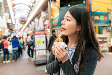 closeup shot of asian woman visitor having baked doughnut and looking into space trying to find her way out at the exit of kuromon ichiba market in Osaka japan