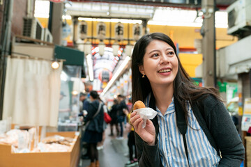 cheerful asian girl backpacker holding eating a donut is having fun visiting kuromon ichiba market in Osaka japan. she looks into space with smile on face.