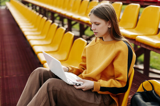 A young girl is doing her homework using a laptop in the stands of the stadium. The concept of training and education