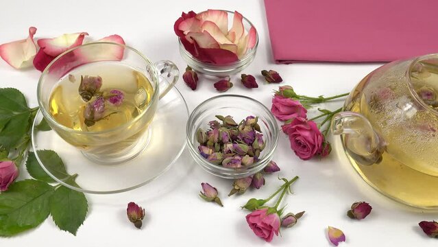 Pink dried rose buds flower tea in a glass teapot and in cup, fresh pink roses, rose petals and dried rose buds are on a white table