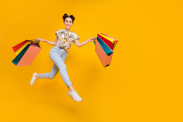 Fototapeta na wymiar Photo of funky excited woman wear print t-shirt jumping high walking holding shoppers empty space isolated yellow color background