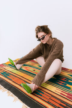 Portrait of stylish young girl in trendy sunglasses, oversized sweater and white tights sitting on vintage carpet isolated over grey studio background