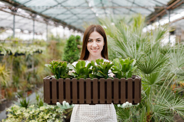 Portrait of a hardworking european farmer woman in a greenhouse holding box with flower pots. Home gardening, love of plants and care. Small business.