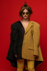 Stylish portrait of young girl wearing two oversized jackets, stylish extravagant glasses, posing isolated over red background