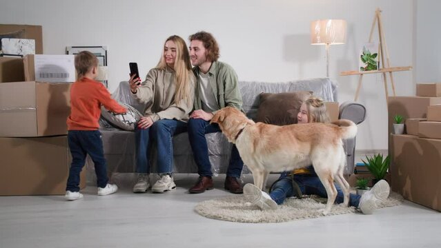 husband and wife sitting on the couch covered with polyethylene talking online using mobile phone, family with children and a labrador dog rejoice at a housewarming