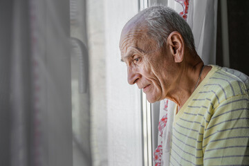 Lonely granddad looking through window glass standing at home. Old man waiting visitors with hope.