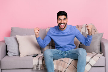 Photo of funny delighted handsome guy sit couch raise fists celebrate success triumph isolated on pink color background
