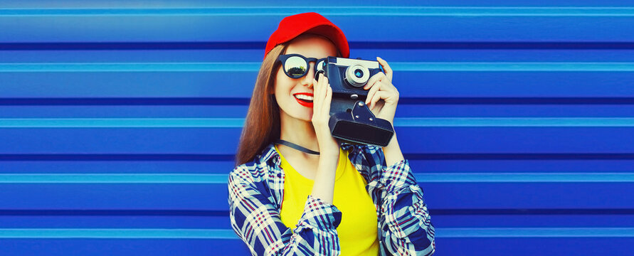 Portrait of happy young woman photographer taking picture on film camera wearing red baseball cap on blue background, banner blank copy space for advertising text